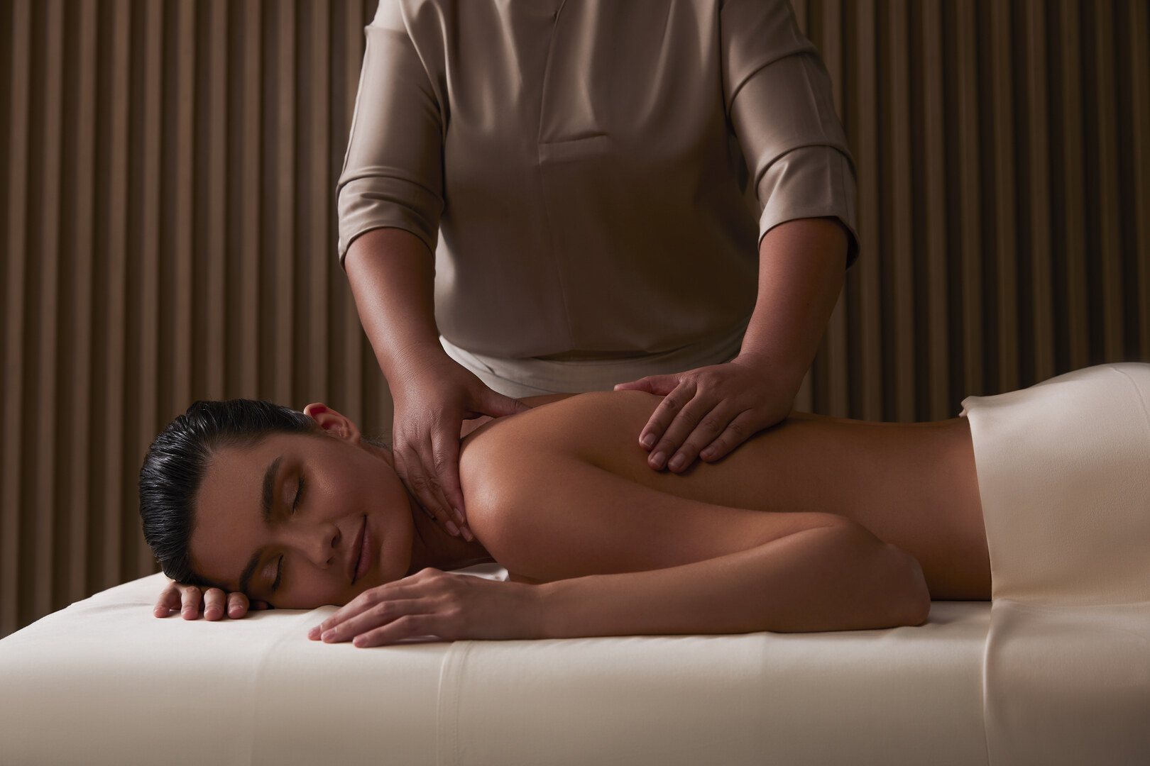 Serene spa setting with a woman lying on a massage table receiving a relaxing back and shoulder massage from a therapist. The tranquil ambiance is enhanced by soft lighting and a warm, neutral-toned interior, creating a calming wellness experience.